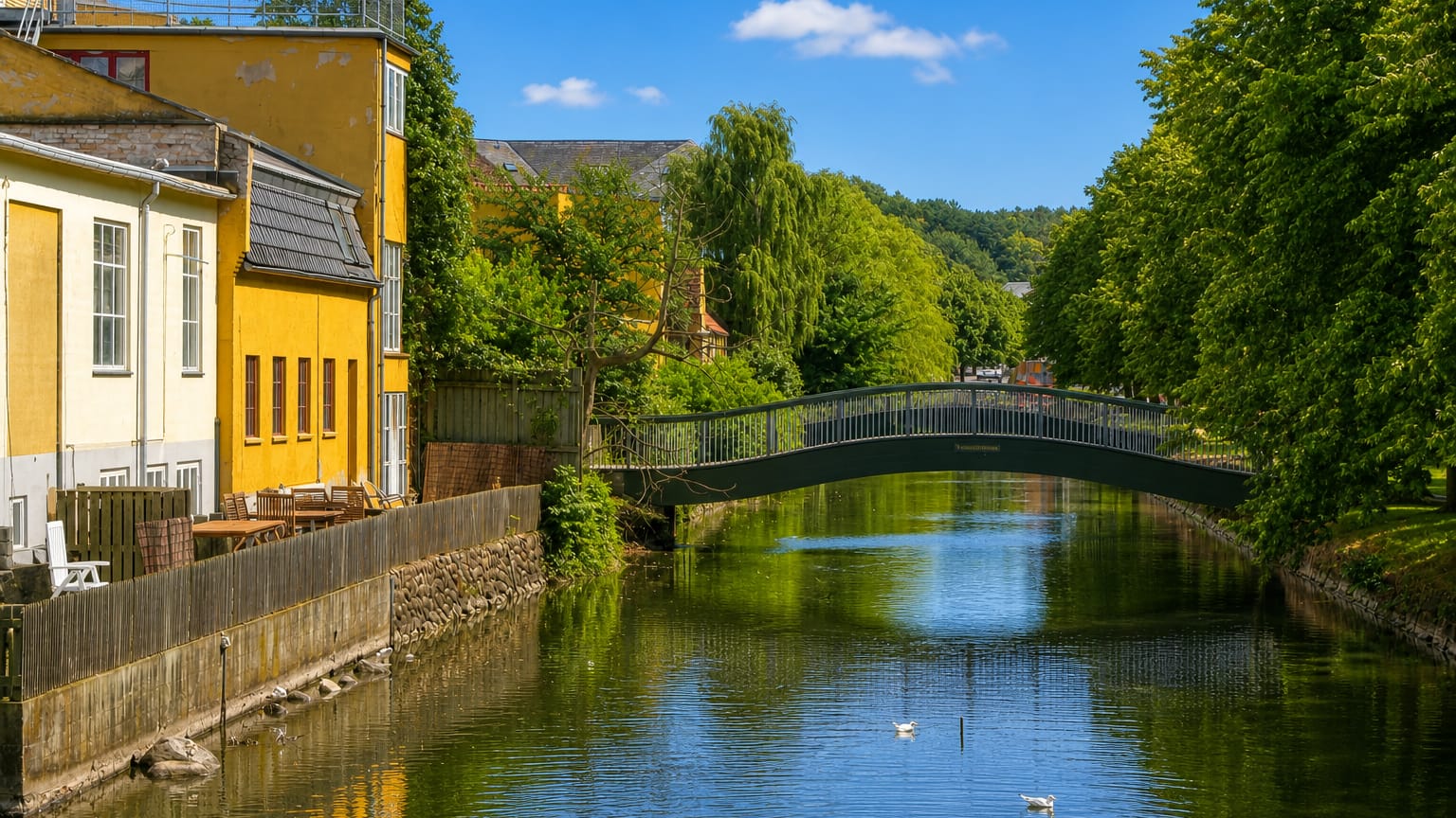 A green arched footbridge over the canal in Frederiksvaerk, Denmark, with a yellow building and swans on the water.