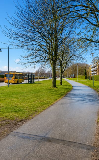Cycle path beside yellow apartment blocks and a bus stop in suburban Glostrup, Denmark, under a clear spring sky.