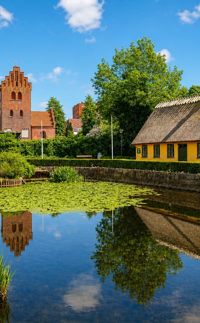Yellow thatched cottage and red-brick stepped-gable church reflected in a pond in Herlev, Denmark
