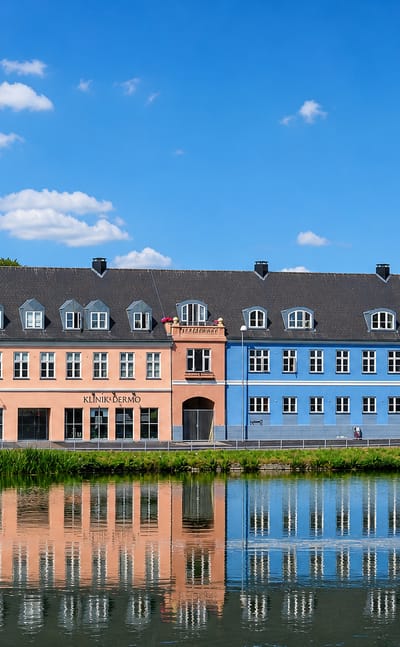 Colourful Hillerod townhouses reflected in a lake, with shop signs including Slotsfrisoren under a blue sky.