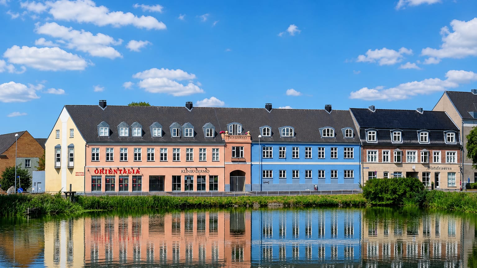 Colourful Hillerod townhouses reflected in a lake, with shop signs including Slotsfrisoren under a blue sky.