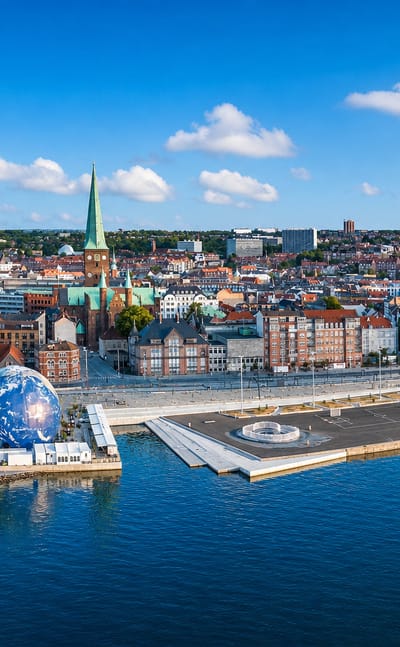 Aerial view of Aarhus C waterfront with the cathedral's green spire, red-tiled rooftops and bay under a blue sky.