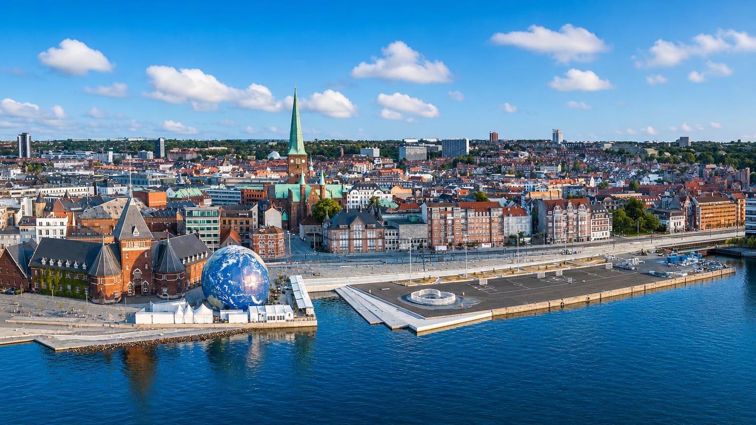 Aerial view of Aarhus C waterfront with the cathedral's green spire, red-tiled rooftops and bay under a blue sky.