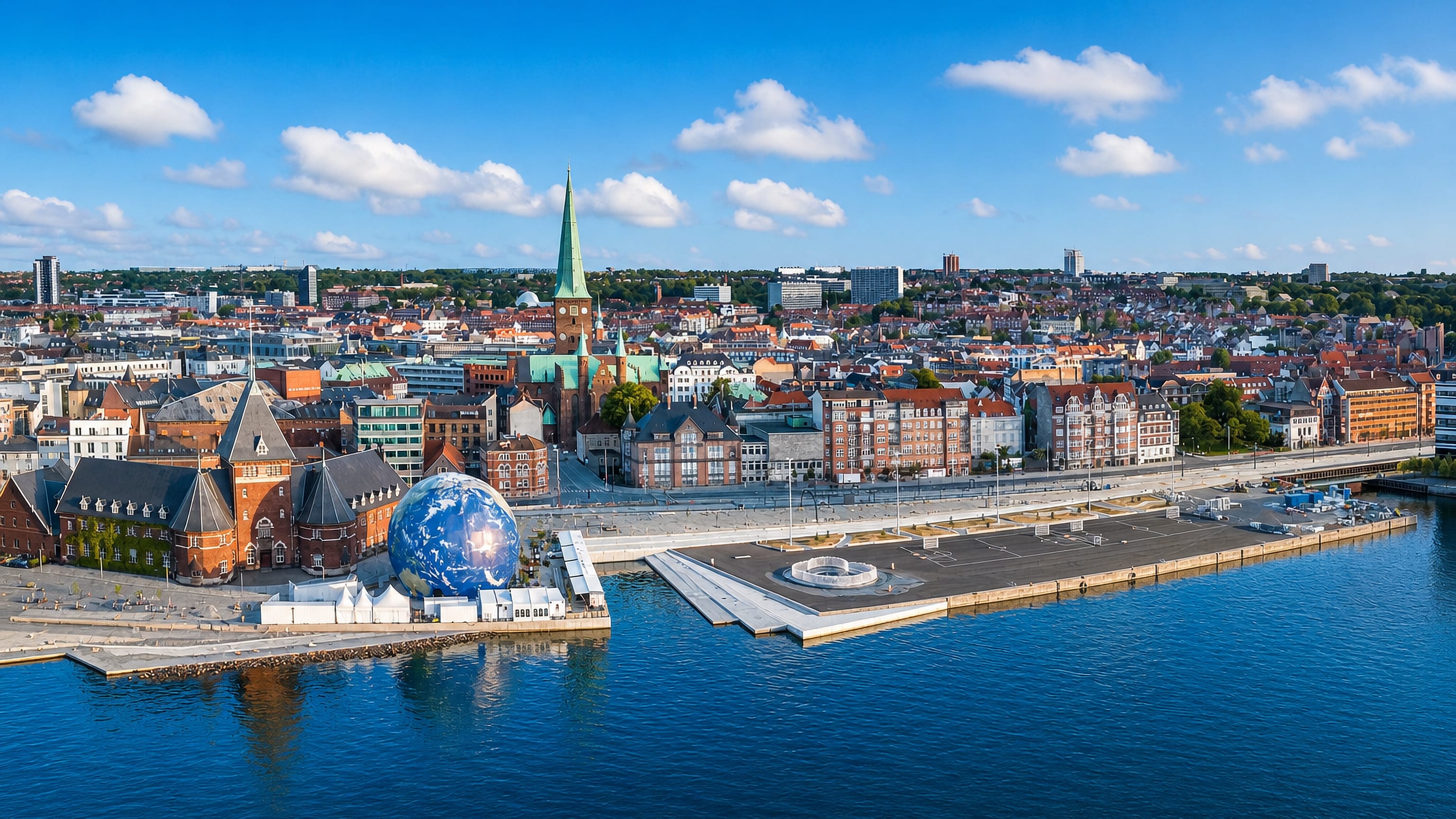 Aerial view of Aarhus C waterfront with the cathedral's green spire, red-tiled rooftops and bay under a blue sky.