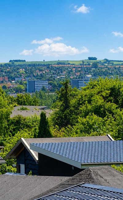Leafy suburban rooftops and a Danish flag in Aarhus V, with distant high-rise housing blocks and green hills under a blue summer sky.