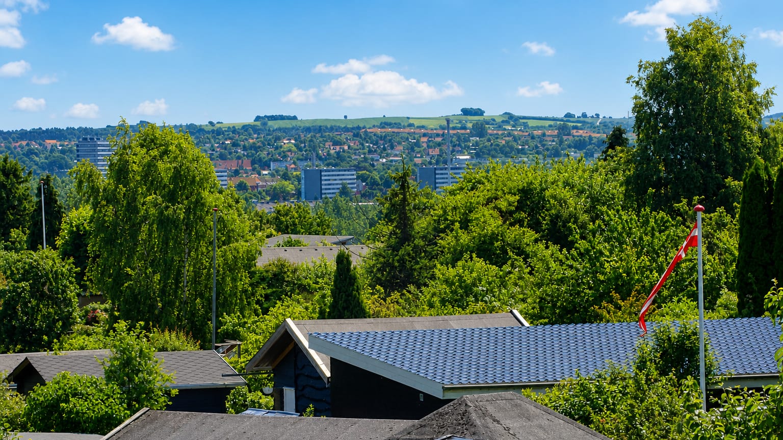 Leafy suburban rooftops and a Danish flag in Aarhus V, with distant high-rise housing blocks and green hills under a blue summer sky.