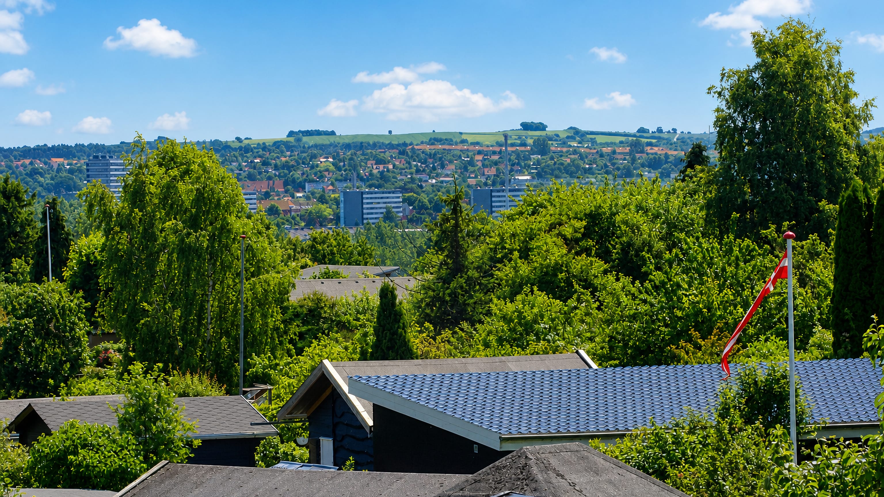 Leafy suburban rooftops and a Danish flag in Aarhus V, with distant high-rise housing blocks and green hills under a blue summer sky.