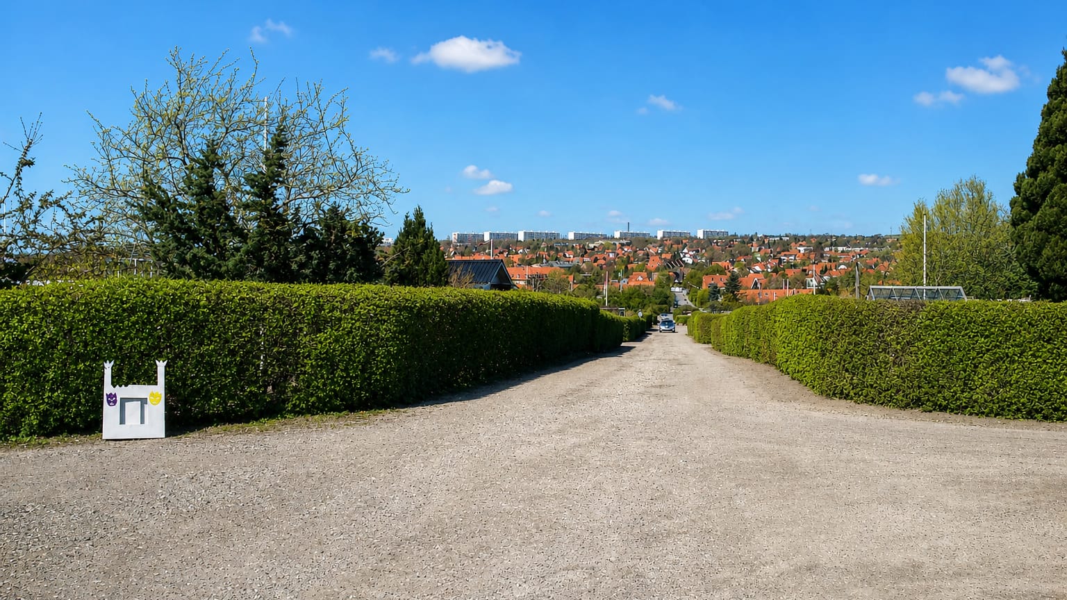 Gravel path between trimmed hedges opening onto Åbyhøj's red-roofed houses and distant apartment blocks under a blue sky