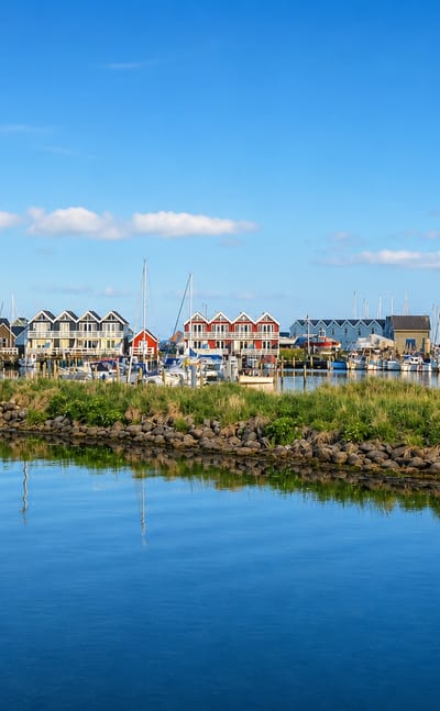 Grenaa harbour on a sunny day, with colourful gabled townhouses, a yacht marina and a rocky breakwater reflected in calm blue water.