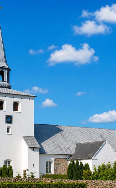 Whitewashed Danish church with a tall lead spire behind a fieldstone dyke under blue sky in Hedensted.