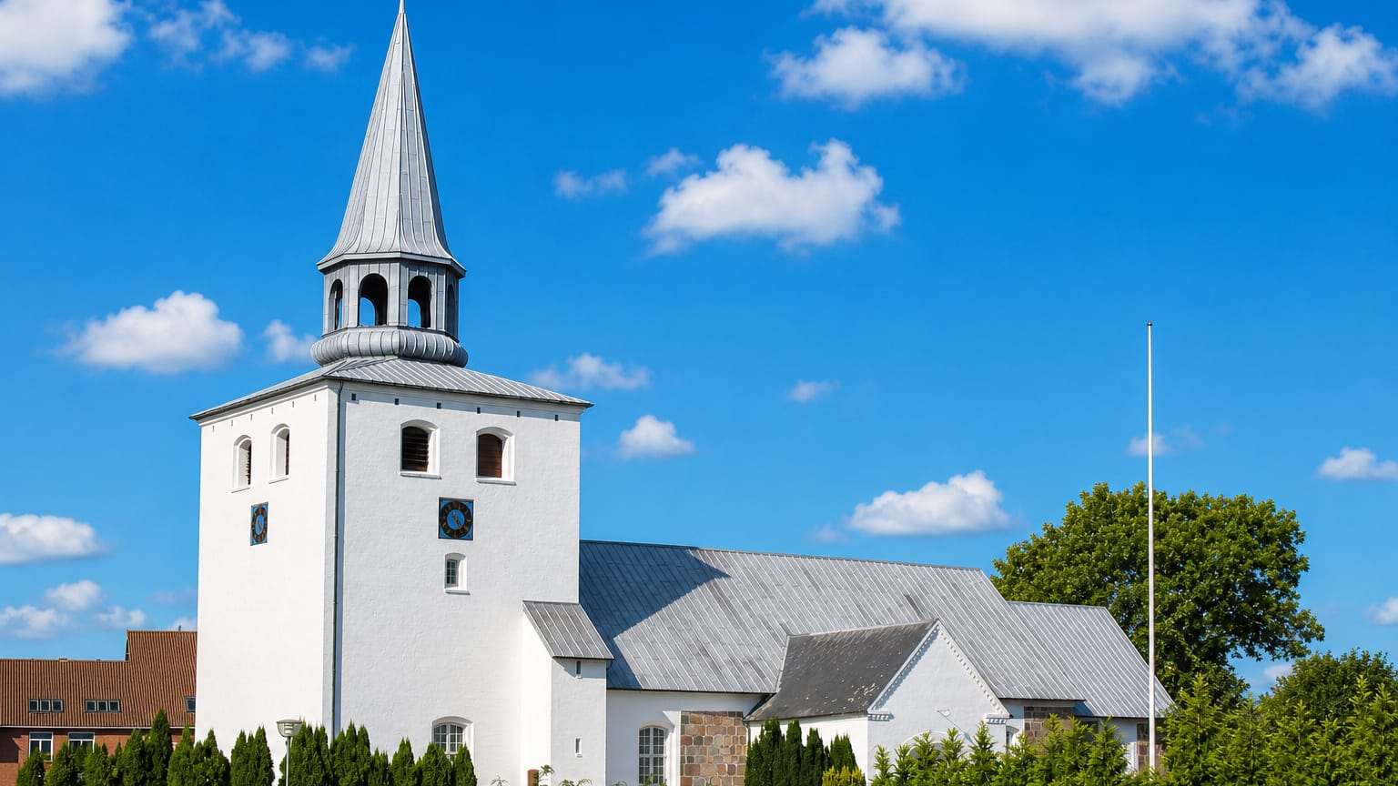 Whitewashed Danish church with a tall lead spire behind a fieldstone dyke under blue sky in Hedensted.