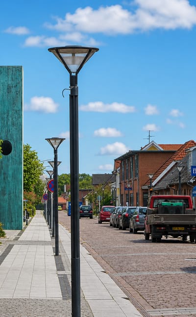 Danish high street with shops, a tall patinated sculpture and parked cars on a paved road in Hinnerup.