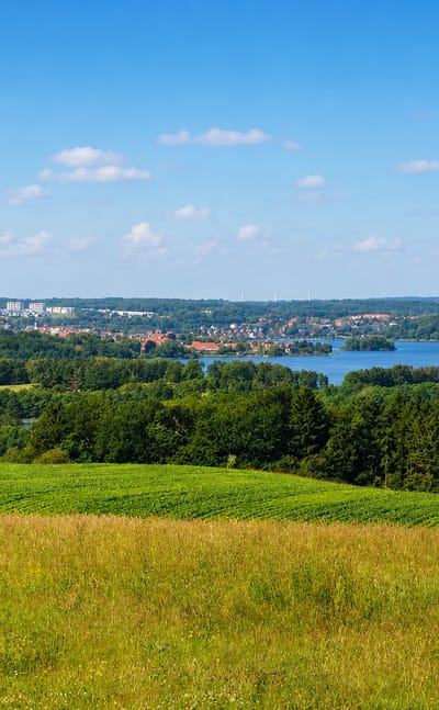 Distant view of Skanderborg, Denmark, beside its lake, seen across rolling green farm fields and woodland under a bright sky.