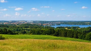 Distant view of Skanderborg, Denmark, beside its lake, seen across rolling green farm fields and woodland under a bright sky.