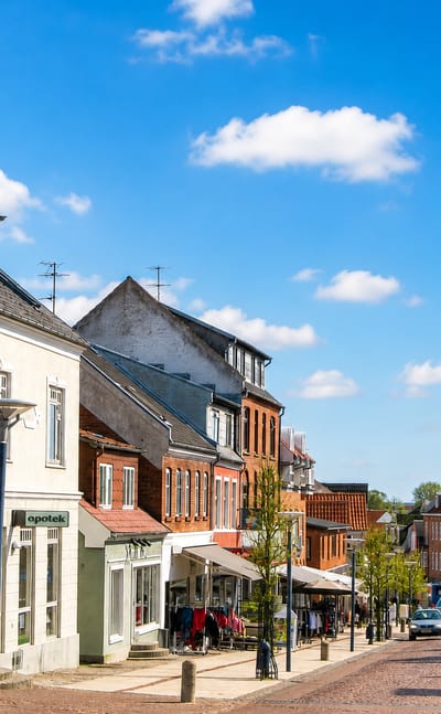 Town-centre street with a white spired pharmacy building under a blue sky in Aars, Denmark.
