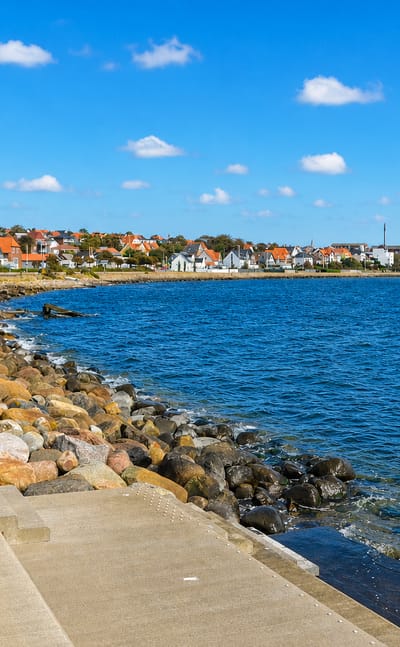 Thisted, Denmark, along its rocky fjord-side promenade, with red-roofed houses lining the distant shore under a blue sky.