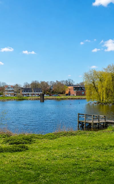 Lakeside view of Kirke Hvalsø, Denmark, with a small wooden jetty, grassy bank, and houses among trees across the water.
