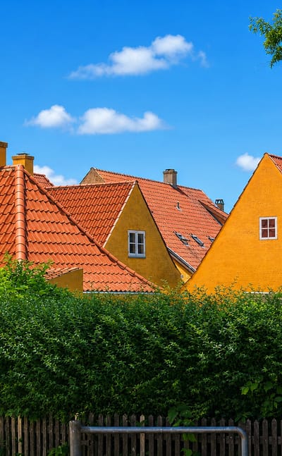 Red-tiled roofs of yellow gabled houses rising above a hedge and picket fence in Maribo, Denmark.