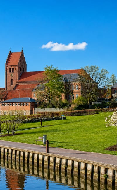 Næstved's red-brick medieval church and old town beside a grassy canal-side promenade in spring