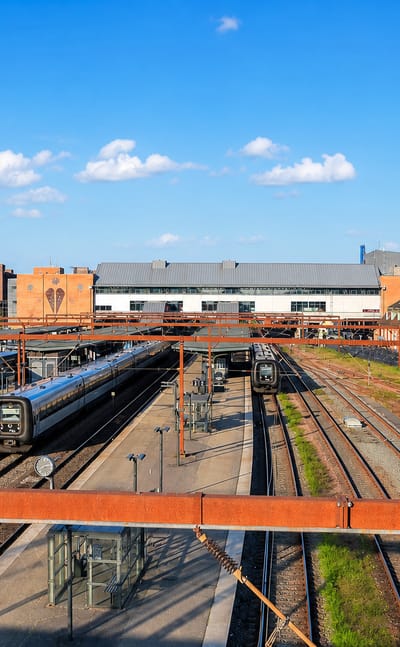Odense railway station with commuter trains, tracks and city rooftops under a bright blue Danish sky.
