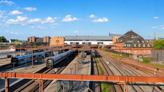 Odense railway station with commuter trains, tracks and city rooftops under a bright blue Danish sky.