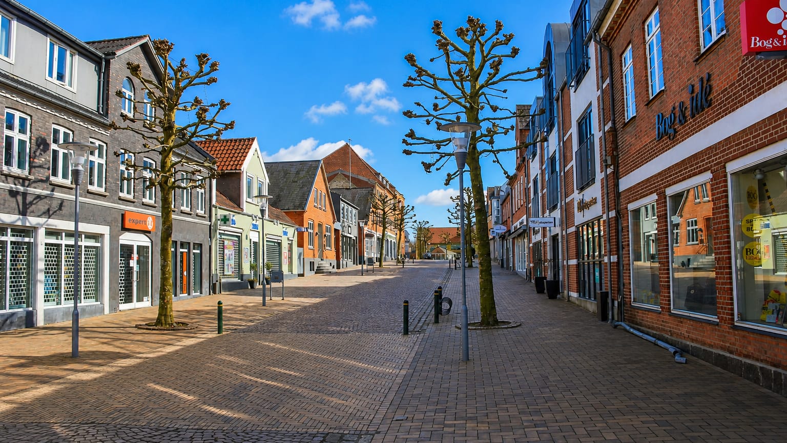 A cobbled pedestrian shopping street lined with brick shops and pollarded trees in central Ringe, Denmark.