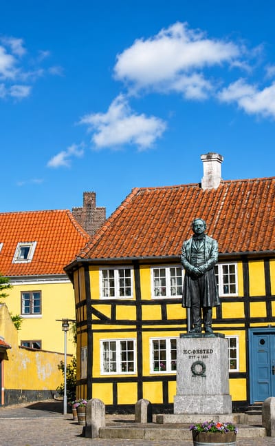 The H.C. Orsted statue before a yellow half-timbered house and church spire in Rudkobing, Denmark.