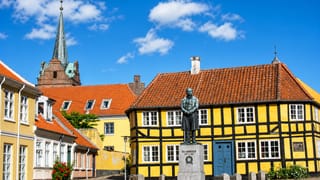 The H.C. Orsted statue before a yellow half-timbered house and church spire in Rudkobing, Denmark.