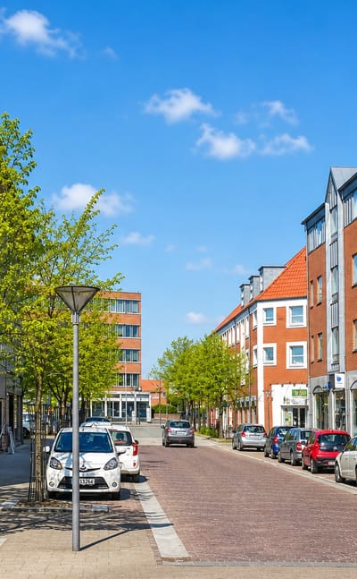 Vejen's modern pedestrian street with glass and red-brick buildings, young trees and parked cars in summer.