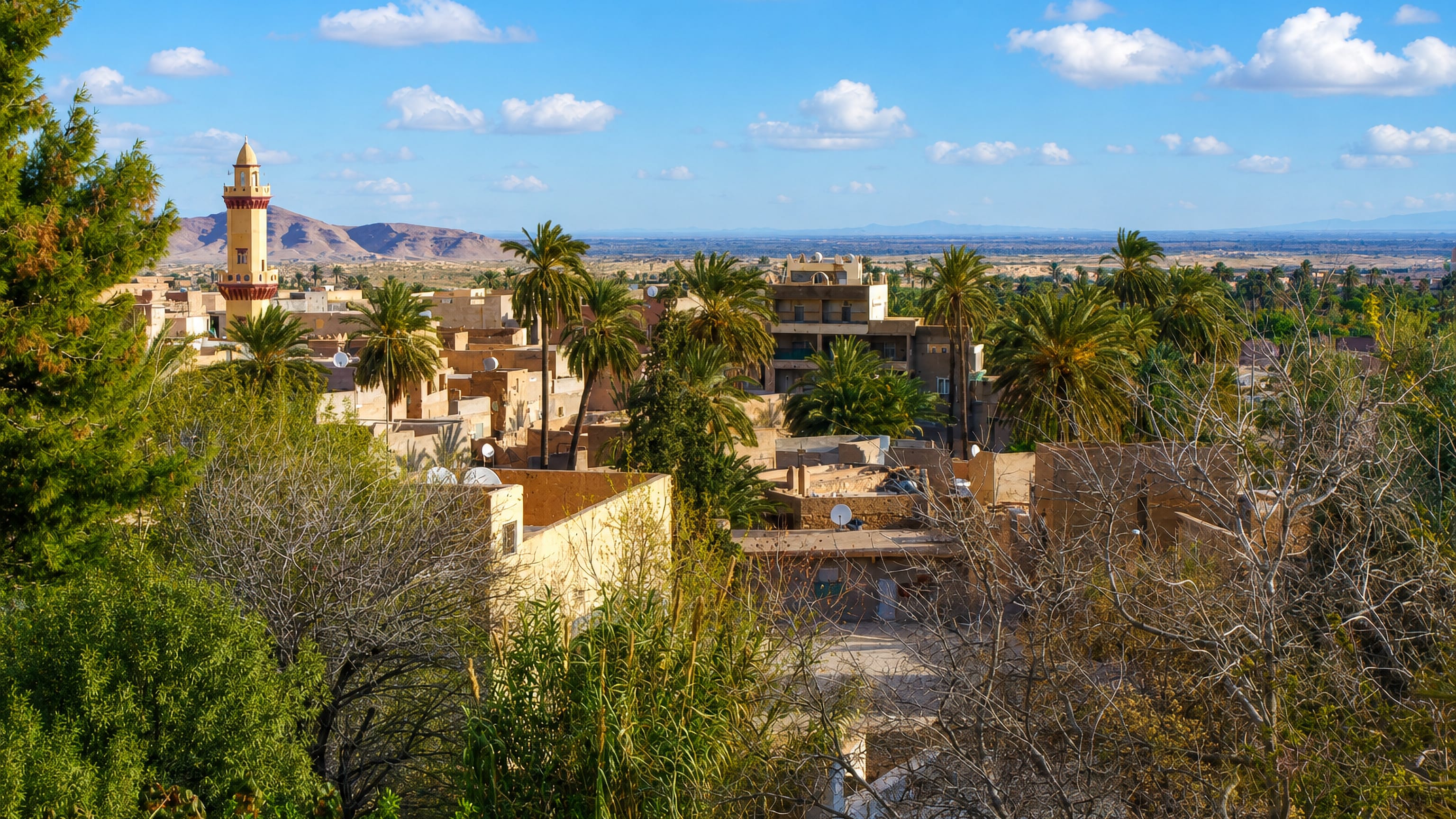 Palm groves and ochre mud-brick houses around a minaret in Bou Saada, Algeria, with arid hills beyond