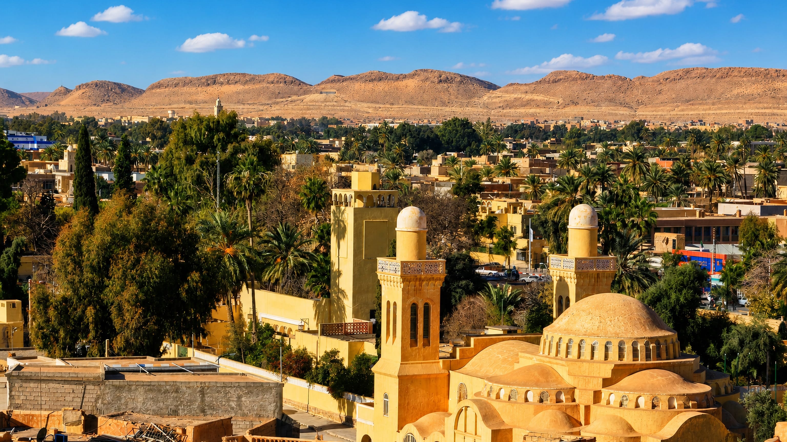 Ochre domes and minarets rising among palms in Laghouat, Algeria, with Saharan Atlas ridges behind.