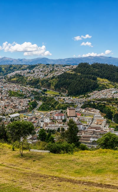 Quito spread across Andean hillsides and valleys beneath mountain ranges, seen from a grassy ridge