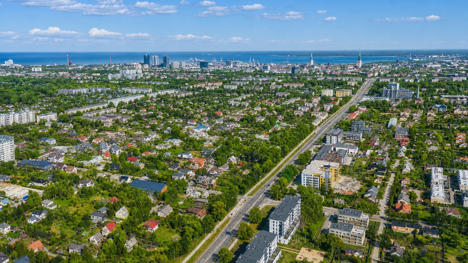 Aerial daytime view of Kristiine in Tallinn, leafy low-rise streets in front with the city centre spires, high-rises and sea beyond.