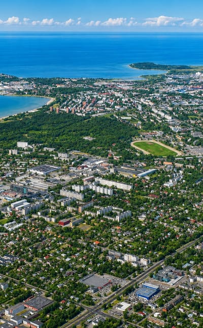 Aerial daytime view of Põhja-Tallinn, a coastal district with a bay, beach, port peninsula and dense urban sprawl under a blue sky.