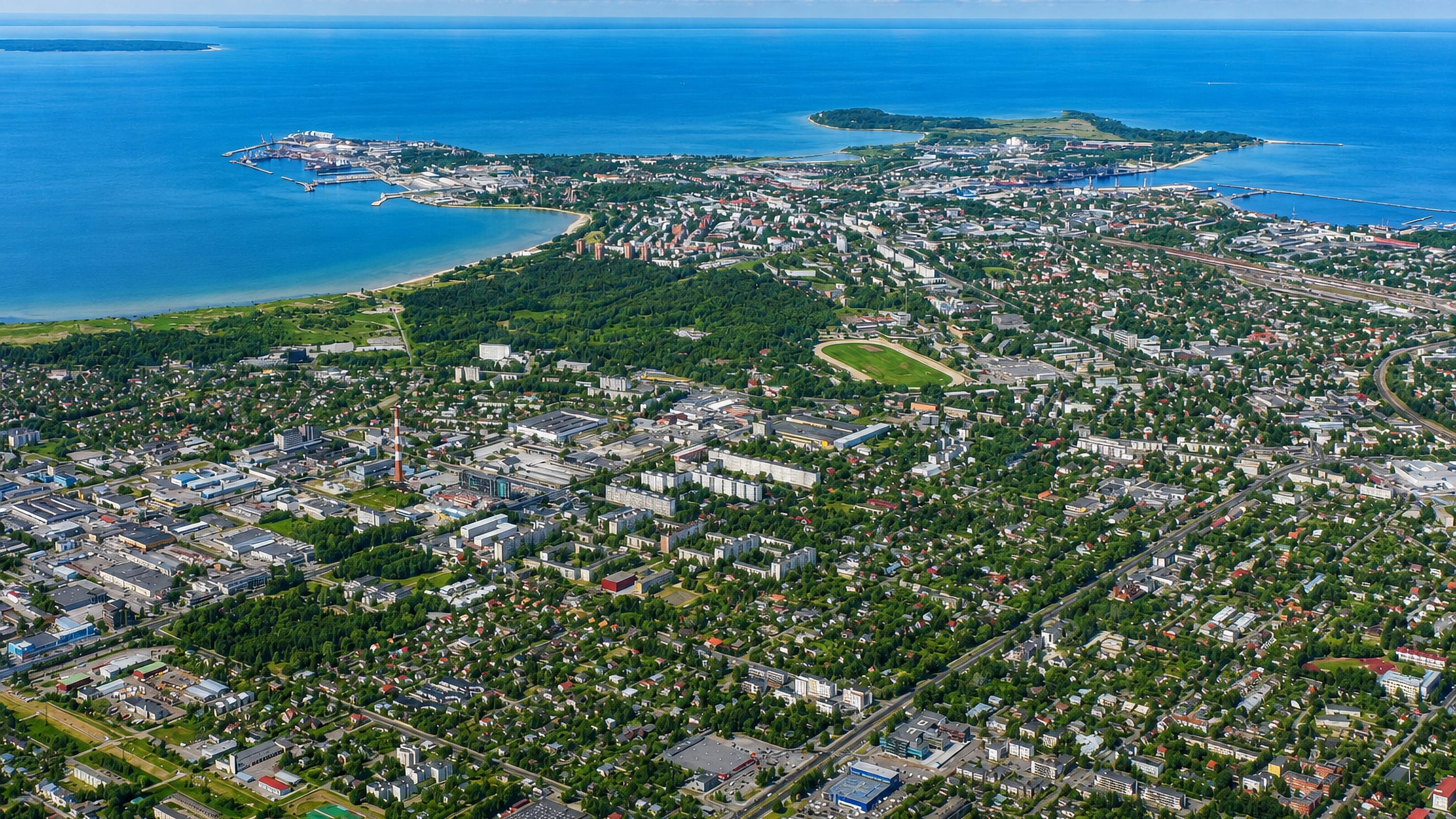 Aerial daytime view of Põhja-Tallinn, a coastal district with a bay, beach, port peninsula and dense urban sprawl under a blue sky.