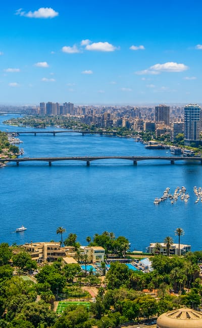 The Nile winding through central Cairo with bridges, river boats and a riverside tower near Hadā'iq al Qubbah.