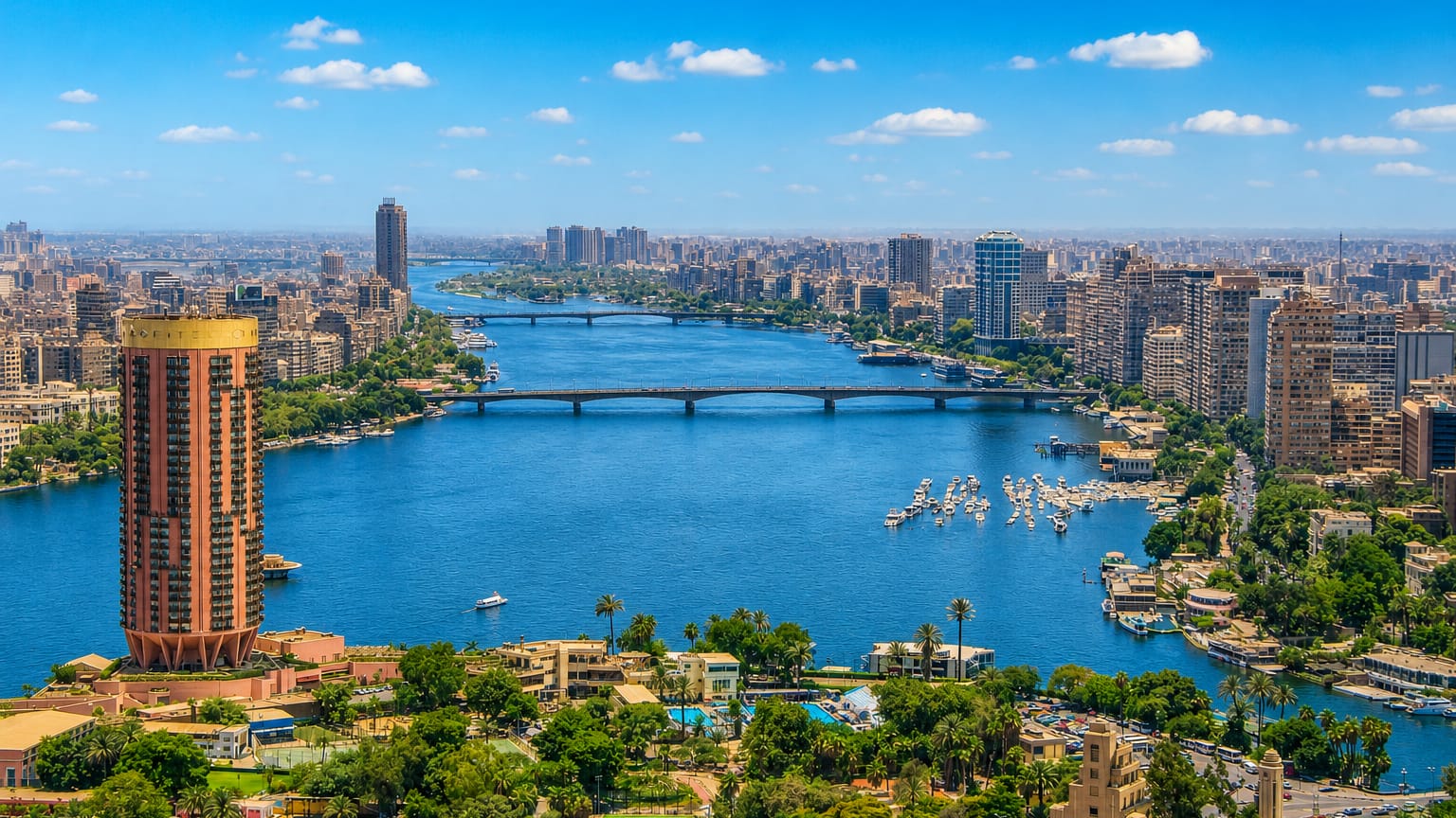 The Nile winding through central Cairo with bridges, river boats and a riverside tower near Hadā'iq al Qubbah.