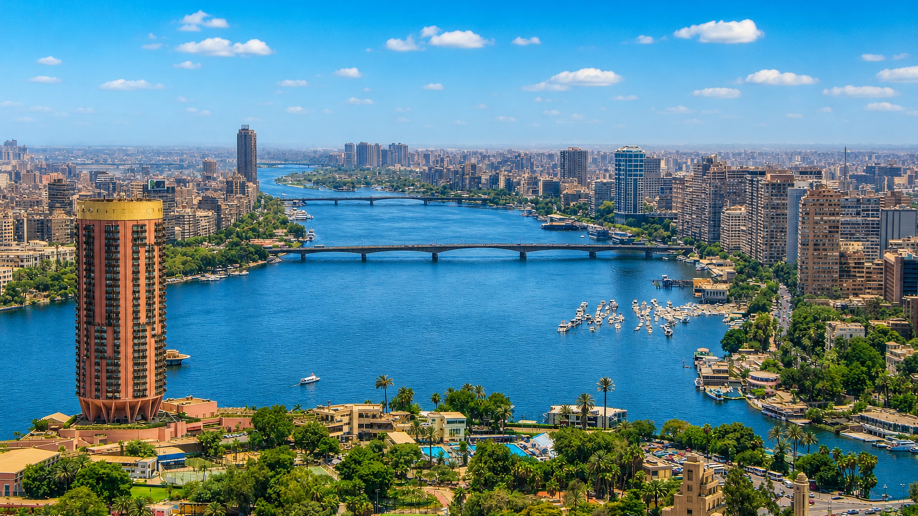 The Nile winding through central Cairo with bridges, river boats and a riverside tower near Hadā'iq al Qubbah.