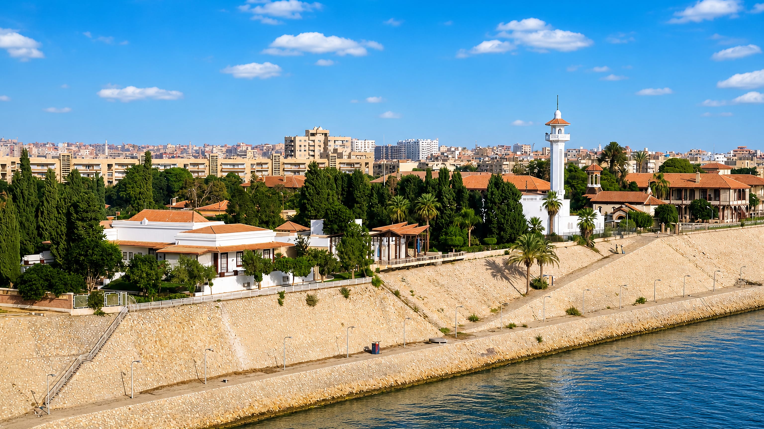 Villas and palms along the Suez Canal embankment at Ismailia, with a white minaret above the waterfront.