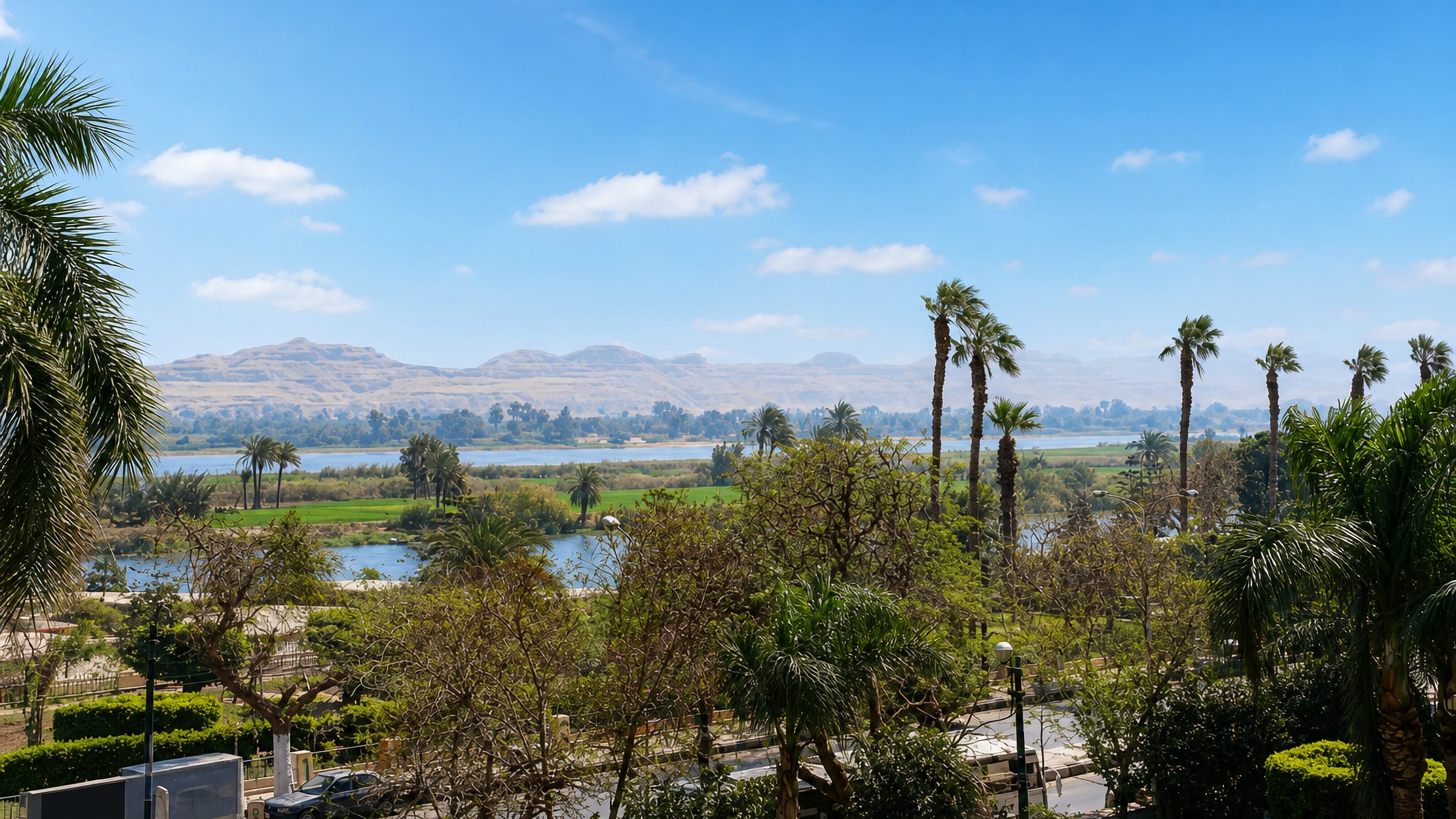 The Nile at Minya seen past palm-lined gardens, with green fields and desert cliffs beyond.