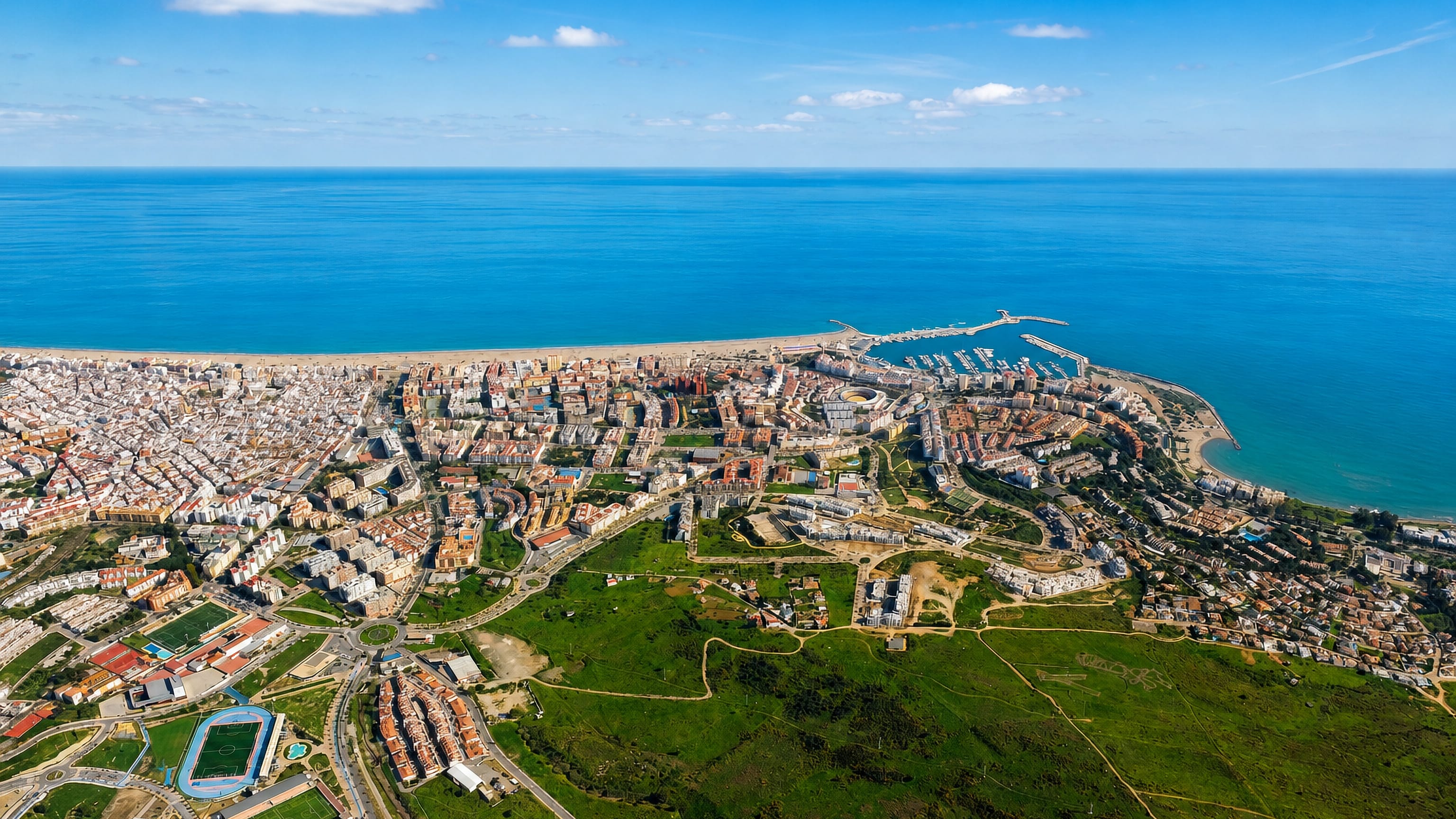 Aerial view of Estepona's whitewashed coastal town, marina and long Mediterranean beach backed by green hills.
