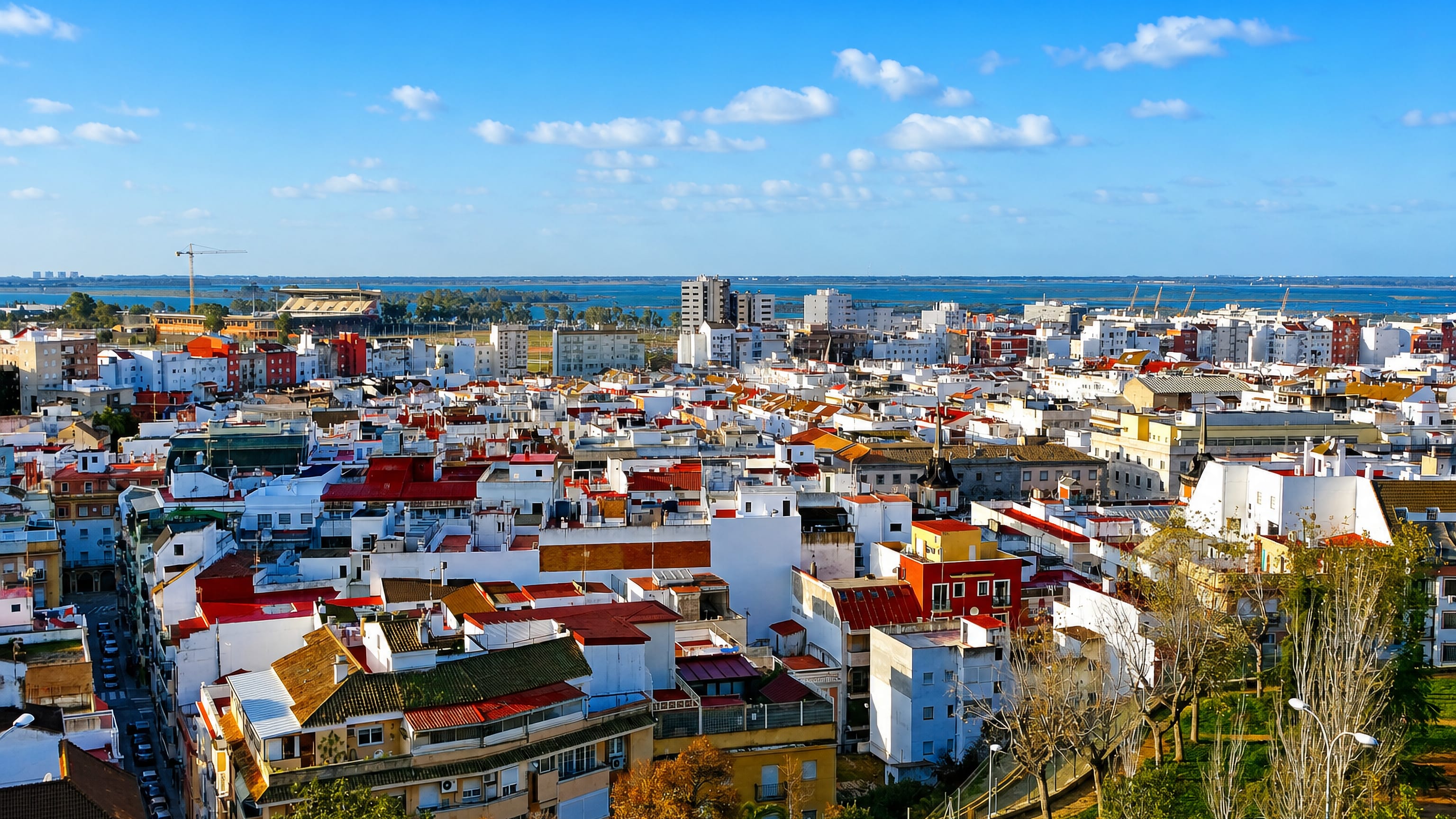 Whitewashed rooftops of Huelva, Spain, spreading toward the Odiel estuary and port cranes under a blue sky.
