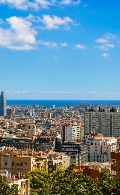Elevated panorama over Barcelona rooftops toward the sea with Torre Glories and the Sagrada Familia on the skyline