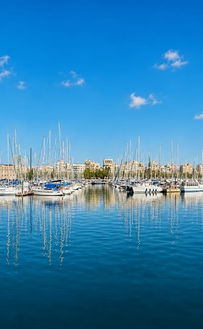 Port Vell marina with moored yachts and the old-town skyline of Ciutat Vella, Barcelona under a bright blue sky.