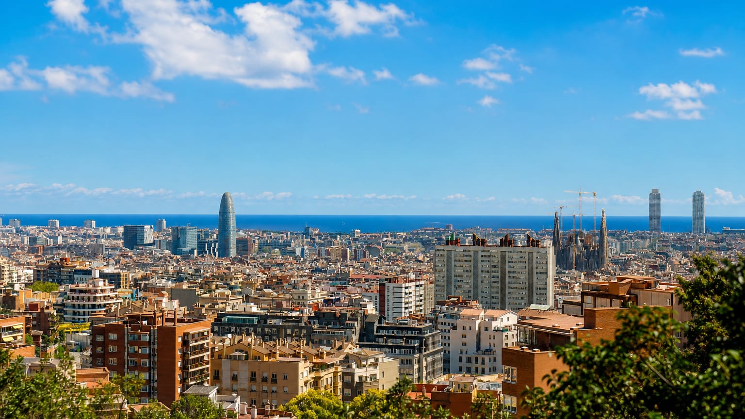 Elevated panorama over Barcelona rooftops toward the sea with Torre Glories and the Sagrada Familia on the skyline
