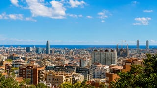 Elevated panorama over Barcelona rooftops toward the sea with Torre Glories and the Sagrada Familia on the skyline