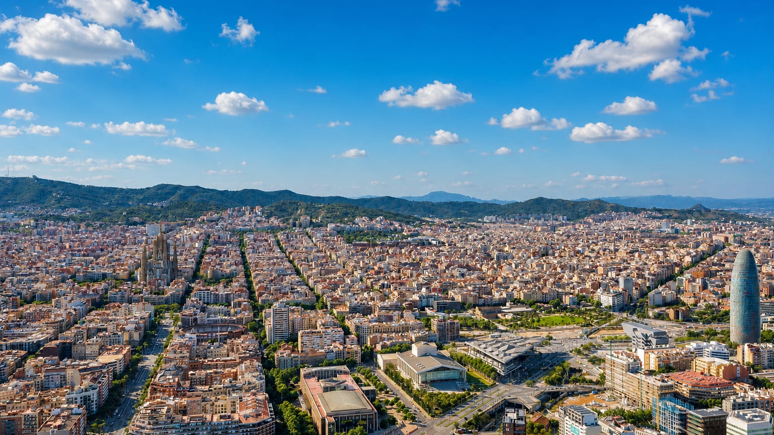 Aerial over Barcelona's Eixample grid with the Sagrada Familia basilica and Torre Glories on the horizon.