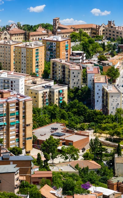Aerial view over the leafy hillside residential district of Sant Genis dels Agudells in Barcelona, with mid-rise blocks among greenery under a blue sky.