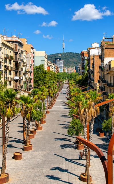 Palm-lined pedestrian rambla in the Sants-Badal district of Barcelona, with the Torre de Collserola rising on the wooded ridge beyond.