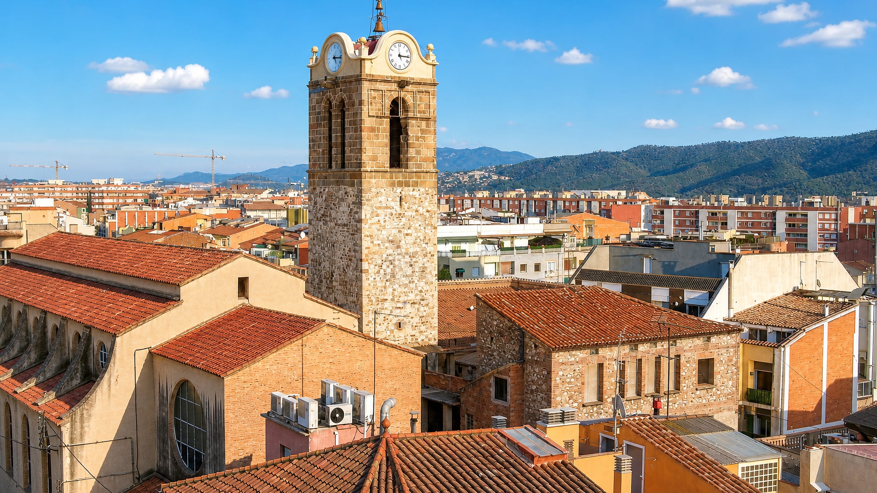 Stone church bell tower with clock faces above tiled rooftops and the hills behind Mollet del Vallès.